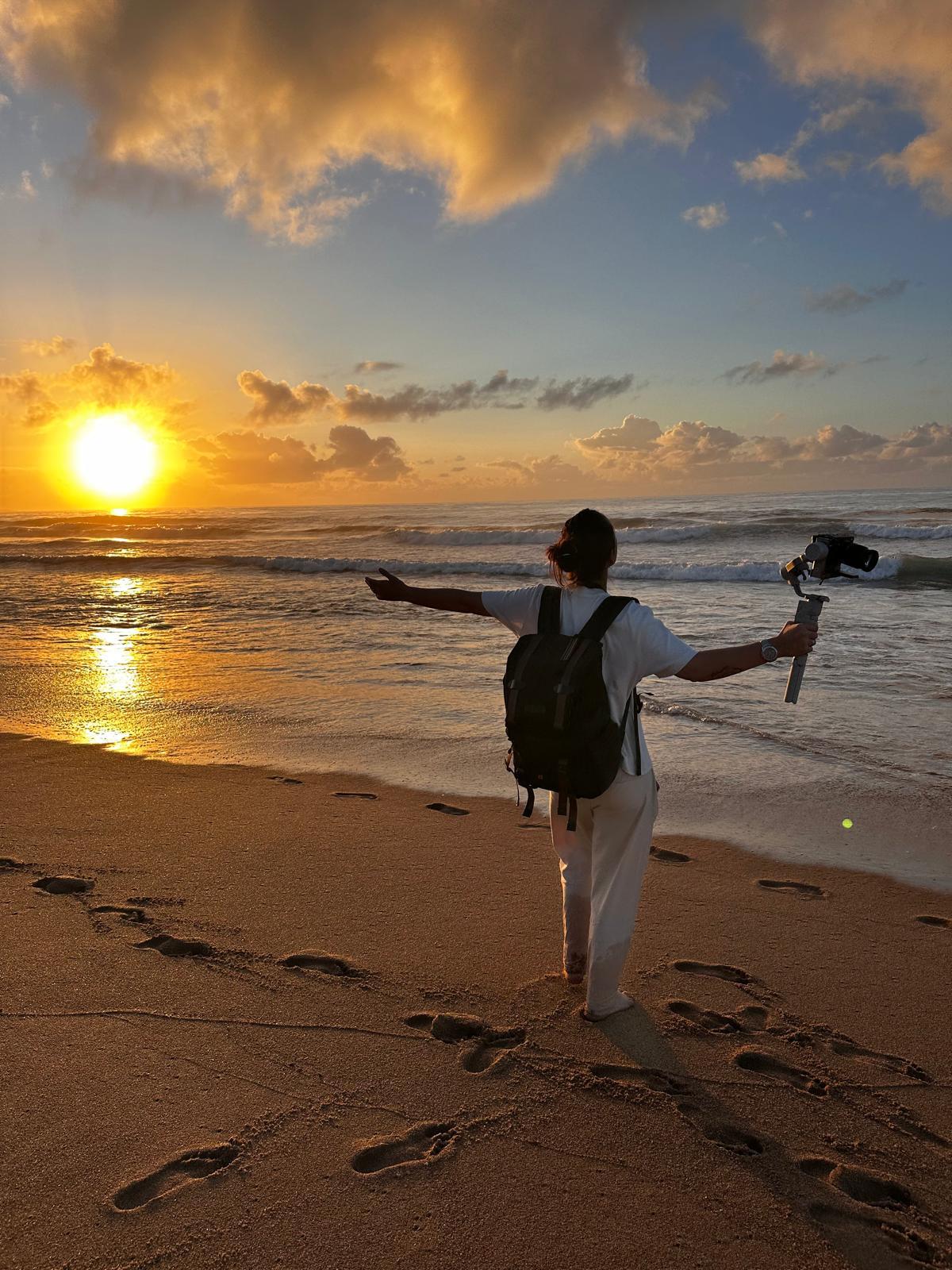 Regiane Severini de braços abertos diante do pôr do sol na praia, com equipamento de filmagem em mãos